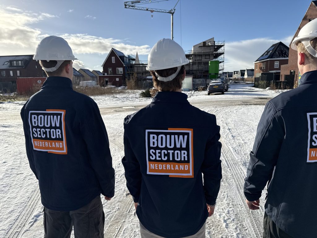Drie vakmensen van Bouwsector Nederland met witte helmen kijken naar een nieuwbouwwijk in de sneeuw